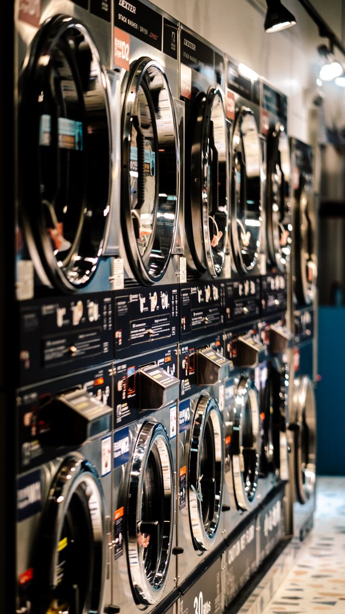 services-01 A row of industrial washing machines in a modern laundromat with vibrant lighting.