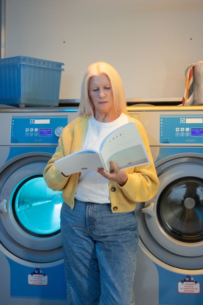 Elderly woman reading a magazine while leaning on a washing machine in a laundry facility.